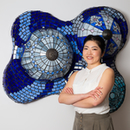 Portrait of Fum Amano in a sleeveless white shirt in front of a sculpture hung on wall, made of various blue glass pieces