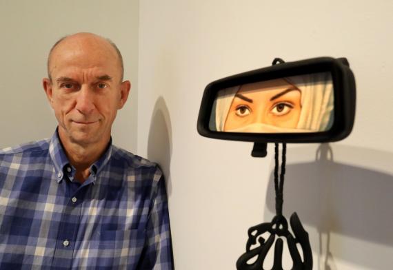 Greg Kucera in blue gingham button-up shirt standing next to an artwork by Humaira Abid. Artwork is of a car rearview window with the reflection of a woman in a cream-colored hijab