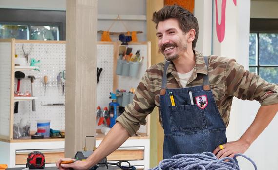 Adam Kingman in a shop, wearing a camouflage button-up shirt with a denim apron