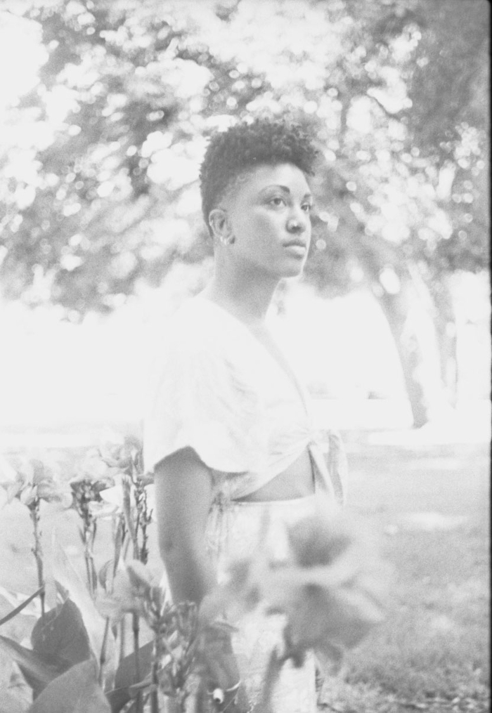 Faded black and white image of Lauren Williams standing near a garden, looking off in the distance, and wearing a cropped short-sleeved shirt