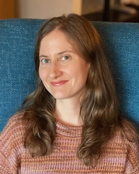 Portrait of Adair Rounthwaite wearing an earth-toned sweater and sitting on a teal colored chair
