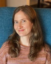 Portrait of Adair Rounthwaite wearing an earth-toned sweater and sitting on a teal colored chair