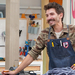 Adam Kingman in a shop, wearing a camouflage button-up shirt with a denim apron