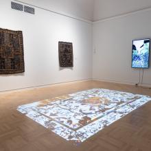 Exhibition view of 2025 MFA + MDes Thesis Exhibition in a darkened room with digital art projected on the floor and tapestries hung on one wall