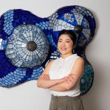 Portrait of Fum Amano in a sleeveless white shirt in front of a sculpture hung on wall, made of various blue glass pieces