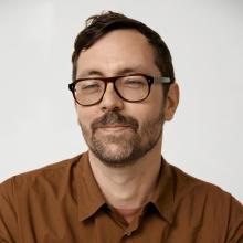 Portrait of Gabriel Stromberg wearing glasses and a burnt orange button up shirt in front of a white background