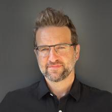 Portrait of Vander McClain wearing glasses and a black button-up shirt in front of a dark grey background