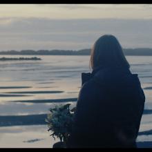 A still shot of a video. There is a figure sitting holding a bouquet of flowers and looking off into a body of water.