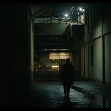 A still shot of a video. There is a figure walking through a dimly-lit alleyway.