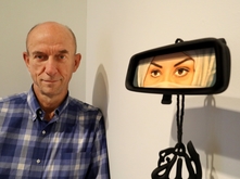 Greg Kucera in blue gingham button-up shirt standing next to an artwork by Humaira Abid. Artwork is of a car rearview window with the reflection of a woman in a cream-colored hijab