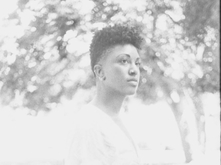 Faded black and white image of Lauren Williams standing near a garden, looking off in the distance, and wearing a cropped short-sleeved shirt