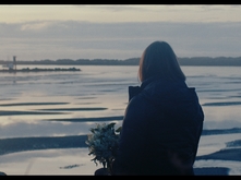 A still shot of a video. There is a figure sitting holding a bouquet of flowers and looking off into a body of water.