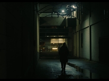 A still shot of a video. There is a figure walking through a dimly-lit alleyway.